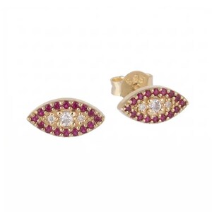 May include: A pair of gold-colored stud earrings in an eye shape. The earrings are adorned with small, round, red gemstones around a central cluster of clear, round gemstones. The earrings have a post and backing.