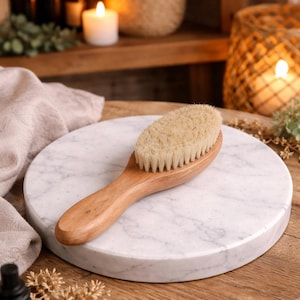 May include: A wooden-handled body brush with natural bristles rests on a white marble surface. The brush is oval-shaped and appears to be made of light-colored wood. The marble surface is round and has gray veining.