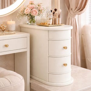 May include: A white vanity with three drawers and gold hardware. A bouquet of pink roses sits in a glass vase on top, along with makeup brushes and perfume bottles. A candle and decorative tray are also visible.