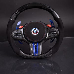 May include: A black and carbon fibre steering wheel with blue and red accents. The wheel features a black suede grip, paddle shifters, and a BMW logo. The centre of the wheel has a blue and red stripe design.