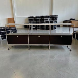 May include: A modern, modular storage unit with a dark brown cabinet and a light-colored top. The unit has a chrome frame and three sections, each with a door and a circular handle. The unit is on a gray concrete floor.