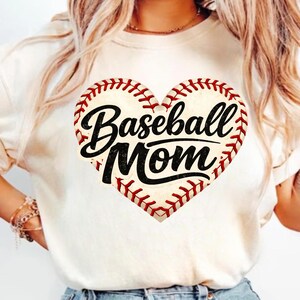 Op de afbeelding: Crèmekleurig T-shirt met een hartvormig honkbalontwerp. Het hart is gestikt met rode draad en heeft de tekst "Baseball Mom" in zwarte letters.