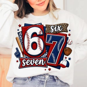 May include: White sweatshirt with a baseball-themed design. Features the numbers "6" and "7" in red, white, and blue, with baseball stitching details. Includes baseball-related graphics and the words "six" and "seven".