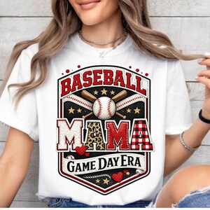 May include: White t-shirt featuring a baseball-themed graphic. The design includes the word "BASEBALL" in red, a baseball, bats, and the word "MAMA" with a leopard print accent. The phrase "GAME DAY ERA" is also included.