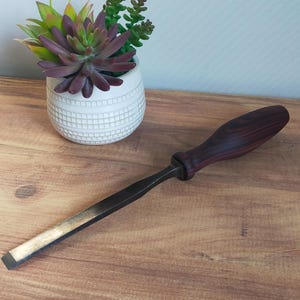 Peut inclure: Un ciseau à bois avec un manche en bois foncé et une lame en métal. Le manche est d'un brun rougeâtre foncé et la lame est gris argenté. Le ciseau est posé sur une surface en bois, avec un petit pot blanc de plantes succulentes en arrière-plan.