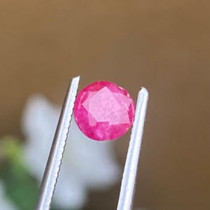 Peut inclure: Une pierre précieuse rubis ronde et facettée, tenue par une pince argentée. Le rubis est d'une couleur rouge-rosé vibrante, avec des facettes visibles qui captent la lumière. L'arrière-plan est un mélange flou de marron et de blanc.