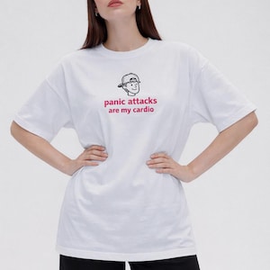 May include: White t-shirt featuring the pink text "panic attacks are my cardio" and a small cartoon graphic. The shirt has a relaxed fit, and the model is wearing black trousers.