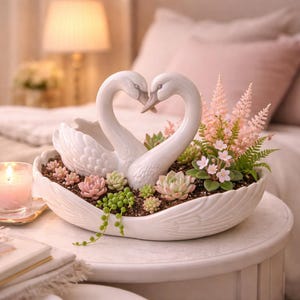 Può includere: Una fioriera a forma di cigno in ceramica bianca con due cigni che formano un cuore. La fioriera è riempita di piante grasse e piante rosa e verdi. Una candela accesa e una lampada sono sullo sfondo.