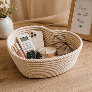 Può includere: Un cesto portaoggetti a forma di cuore realizzato in corda intrecciata color bianco sporco. Il cesto contiene una calcolatrice bianca, uno smartphone di colore chiaro, una chiave dell'auto nera, un mazzo di chiavi, occhiali tondi e due piccoli contenitori circolari.