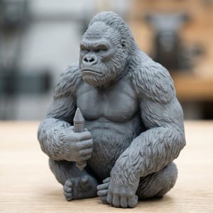 Puede incluir: Una figura gris impresa en 3D de un gorila sentado con complexión musculosa y pelaje texturizado. El gorila sostiene un pequeño objeto puntiagudo con forma de lápiz en su mano derecha. La figura presenta expresiones faciales detalladas.