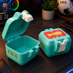 May include: Two square teal storage containers with white plastic latches. One container is open, showing a hollow interior. The other is closed, featuring a red graphic with a gold trophy icon and the text FIFA printed in gold on the side.