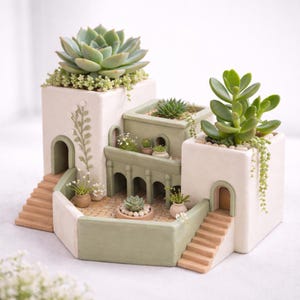 May include: A multi-tiered, white and green ceramic planter shaped like a Mediterranean village. The planter features miniature staircases, archways, and balconies, each holding various succulents and small plants. The overall design is detailed and decorative.