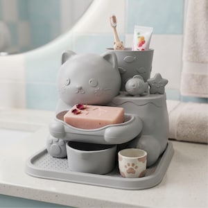 May include: A grey cat-shaped bathroom organiser featuring a soap dish holding a pink bar of soap, a toothbrush holder with a cat-faced toothbrush and toothpaste, a small fish figure, a star figure, and a small cup with a paw print design.