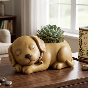 May include: A tan, sleeping puppy-shaped planter with a brown collar and a small gold bell. A green succulent plant is nestled in the opening on the puppy's back. The puppy has closed eyes, a dark brown nose, and a smooth, matte finish.