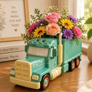 May include: A green ceramic semi-truck planter with the text WILD POT printed on the hood. The truck bed is filled with a colorful floral arrangement featuring pink roses, yellow sunflowers, and purple daisies with green foliage.