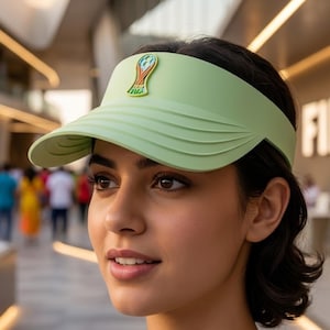 May include: A light green visor featuring a centered emblem with a stylized trophy graphic and the text FIFA. The visor has a smooth, solid-colored brim with subtle horizontal ridges along the sides, providing a clean and functional design.