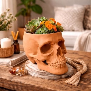 May include: A terracotta skull-shaped planter filled with succulents and orange flowers. The planter sits on a white wooden coaster on a wooden table. Decorative beads and a tassel are on the table.