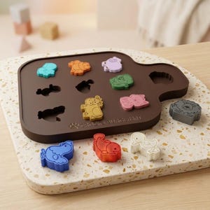 May include: A dark brown silicone mold tray labeled "CROCKS BURGER KIT" with various animal-shaped cutouts. Several colorful silicone animal inserts, including a blue chicken, red fish, white duck, yellow horse, and gray lion, are placed on and around the tray.