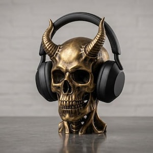 May include: A bronze-coloured skull sculpture featuring two prominent curved horns, serving as a stand for a pair of matte black over-ear headphones. The skull has detailed bone textures and deep eye sockets, with the headphones resting on the cranium.