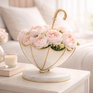 May include: A white and gold umbrella-shaped flower vase filled with pink peonies. The vase sits on a marble base and has a gold handle. A candle and a book are on the table next to the vase.