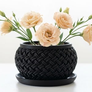 May include: A matte black planter with a textured, woven-style exterior pattern and a matching circular saucer base. The planter contains several pale peach-colored eustoma flowers with green stems and unopened buds extending upwards.