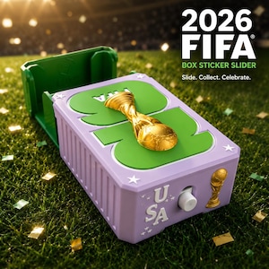May include: A light purple 2026 FIFA box sticker slider featuring a gold trophy emblem on top, a white sliding button, and the text USA with small star accents. The rectangular container has a green interior and a textured side panel design.