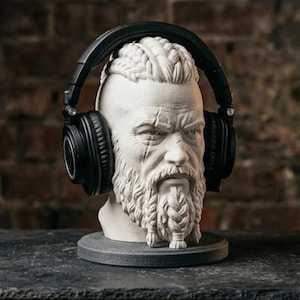 May include: A white stone-like bust of a bearded person with braided hair and a scarred forehead, wearing black over-ear headphones. The sculpture is mounted on a circular base, featuring detailed facial textures and intricate hair braiding.