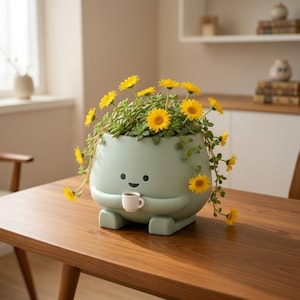 May include: A light green ceramic planter shaped like a character, holding a small white cup. The planter is filled with green succulents and yellow flowers. The planter sits on a wooden table.