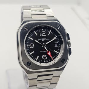 Könnte beinhalten: Eine automatische Bell & Ross-Uhr aus Edelstahl mit quadratischem Zifferblatt. Die Uhr hat ein schwarzes Zifferblatt mit weißen Ziffern und Zeigern sowie einen rotspitzigen Sekundenzeiger. Das Armband ist ebenfalls aus Edelstahl.