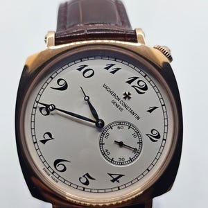 May include: A gold Vacheron Constantin wristwatch with a white face and black numerals. The watch has a small seconds dial and a brown leather strap. The words "Vacheron Constantin Geneve" and "Swiss Made" are visible.