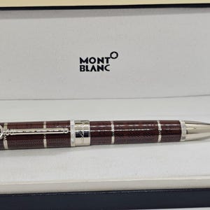 May include: A Montblanc ballpoint pen in a presentation box. The pen features a brown plaid barrel with silver detailing. The pen's tip is silver. The Montblanc logo is displayed on the pen and the box.