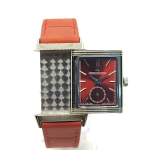 Peut inclure: Une montre-bracelet rectangulaire avec un cadran rouge et un boîtier argenté. La montre a un bracelet en cuir rouge. Le cadran de la montre porte l'inscription "Jaeger-LeCoultre" et un petit cadran des secondes. La montre est exposée sur un support transparent.