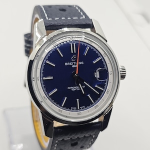 Op de afbeelding: Een Breitling Top Time horloge met een marineblauwe wijzerplaat en zilveren kast. Het horloge heeft een zwarte leren band met witte stiksels. De wijzerplaat heeft witte uurmarkeringen, een datumvenster en een oranje secondewijzer.