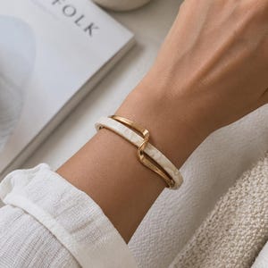Peut inclure: Un bracelet jonc articulé présentant un design à double couche avec une bande en métal doré lisse et une bande en résine marbrée de couleur crème qui se chevauchent en forme de vague, créant une esthétique sophistiquée et superposée.