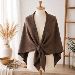 Peut inclure: Un châle marron foncé uni drapé sur un buste de mannequin beige. Le tissu présente un aspect texturé et légèrement duveteux, noué par un simple nœud au centre sur le devant, créant deux extrémités pointues qui pendent sous le nœud.