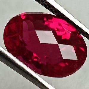 Op de afbeelding: Een gefacetteerde, ovaalvormige robijn edelsteen, vastgehouden door een zilveren pincet. De robijn heeft een diepe, levendige rode kleur, met meerdere facetten die het licht reflecteren. De pincet is gepositioneerd om de vorm en helderheid van de edelsteen te tonen.