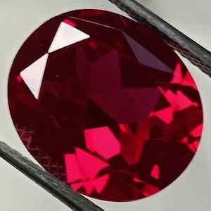 Peut inclure: Un rubis taillé en ovale, maintenu par une pince sur fond blanc. La pierre précieuse rouge vif présente des facettes brillantes. La pince est de couleur gris foncé.