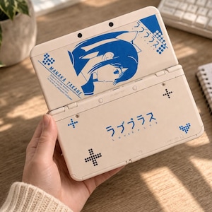 Könnte beinhalten: Eine cremefarbene Handheld-Spielkonsole mit blauen Grafiken und japanischem Text. Die obere Hälfte zeigt eine blaue Illustration im Anime-Stil und den Namen "Manaka Takane". Die untere Hälfte trägt den Text "Love Plus" auf Japanisch. Die Konsole ist geöffnet.