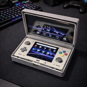 Puede incluir: Consola de juegos portátil en una funda gris, abierta para revelar una pantalla que muestra una interfaz de usuario con iconos de aplicaciones. La consola tiene un pad direccional, palancas analógicas y botones de acción de colores. La hora 3:16 se muestra.