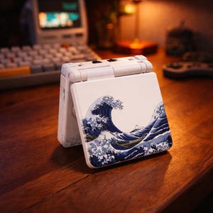 Könnte beinhalten: Eine weiße Handheld-Spielkonsole mit Klappdesign. Die Abdeckung zeigt ein blau-weißes Wellenmuster, inspiriert von japanischer Kunst. Das Gerät ist geöffnet und zeigt den Bildschirm und die Tasten.