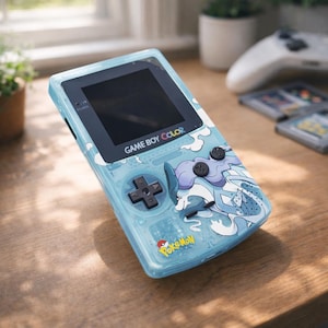 May include: A light blue Game Boy Color handheld console with a Pokemon design. The console features a black screen, directional pad, and buttons. The Pokemon graphic is white and blue. The text "GAME BOY COLOR" and "Pokemon" are visible.