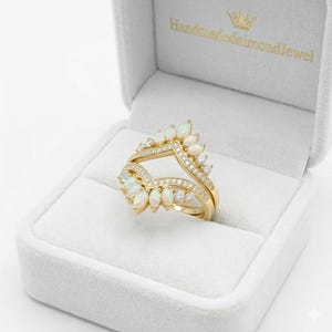 May include: A gold ring featuring a chevron-shaped band adorned with marquise-cut opals and small round clear stones. The ring is displayed inside a light gray velvet jewelry box with the text HandmadeDiamondJewel printed in gold on the lid.