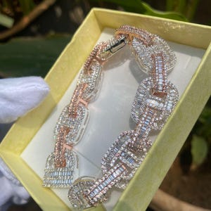 Puede incluir: Un brazalete de oro rosa y plata con piedras rectangulares y baguette. El brazalete se muestra en una caja amarilla, mostrando su intrincado diseño y apariencia brillante. El cierre es visible, destacando la artesanía.