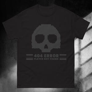 May include: A black short-sleeved t-shirt featuring a dark grey pixelated skull graphic centred on the chest. Below the skull, the text reads 404 ERROR PLAYER NOT FOUND, flanked by two horizontal lines on each side, all in a matching dark grey tone.