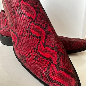Puede incluir: Un par de botines con estampado de serpiente en rojo y negro. Los botines tienen una punta afilada y un tacón bajo negro. El estampado es un diseño llamativo, ideal para añadir un toque especial a cualquier atuendo. Los botines están hechos de un material texturizado.
