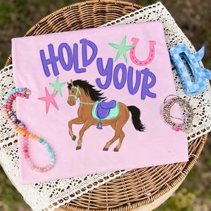 Puede incluir: Una camiseta rosa claro con las palabras "HOLD YOUR" en escritura morada, con un caballo marrón con una silla de montar, estrellas y un gráfico de herradura. La camiseta está rodeada de joyas y un accesorio para el cabello azul.