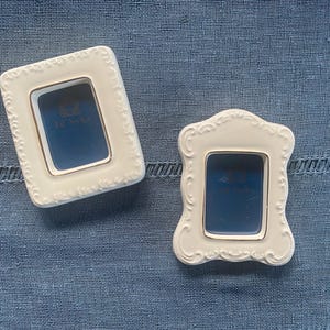 May include: Two white ceramic picture frames with ornate, scrolled edges and gold trim. The frames have a rectangular and a curved shape, both with a blue interior. The frames are on a blue textured surface.