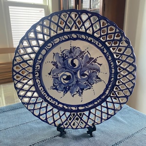 Puede incluir: Un plato decorativo blanco y azul con un diseño floral. El plato presenta un borde calado enrejado y un ramo central de flores azules. Se exhibe sobre un soporte negro, destacando sus intrincados detalles y artesanía.