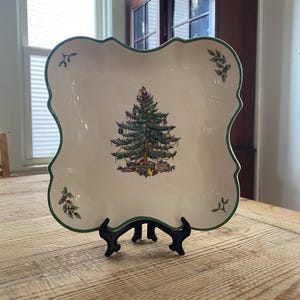 May include: A square, cream-colored ceramic plate with scalloped edges and a green rim. The center features a decorated Christmas tree with gifts underneath. Small sprigs of holly and mistletoe are painted in the corners of the plate.