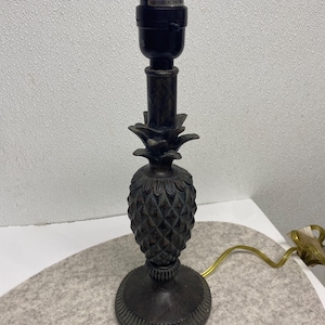 Puede incluir: Una lámpara de mesa de color bronce con una base en forma de piña. La lámpara tiene una base redonda, un cuerpo de piña texturizado y un portalámparas negro. Un cable dorado se extiende desde la base.